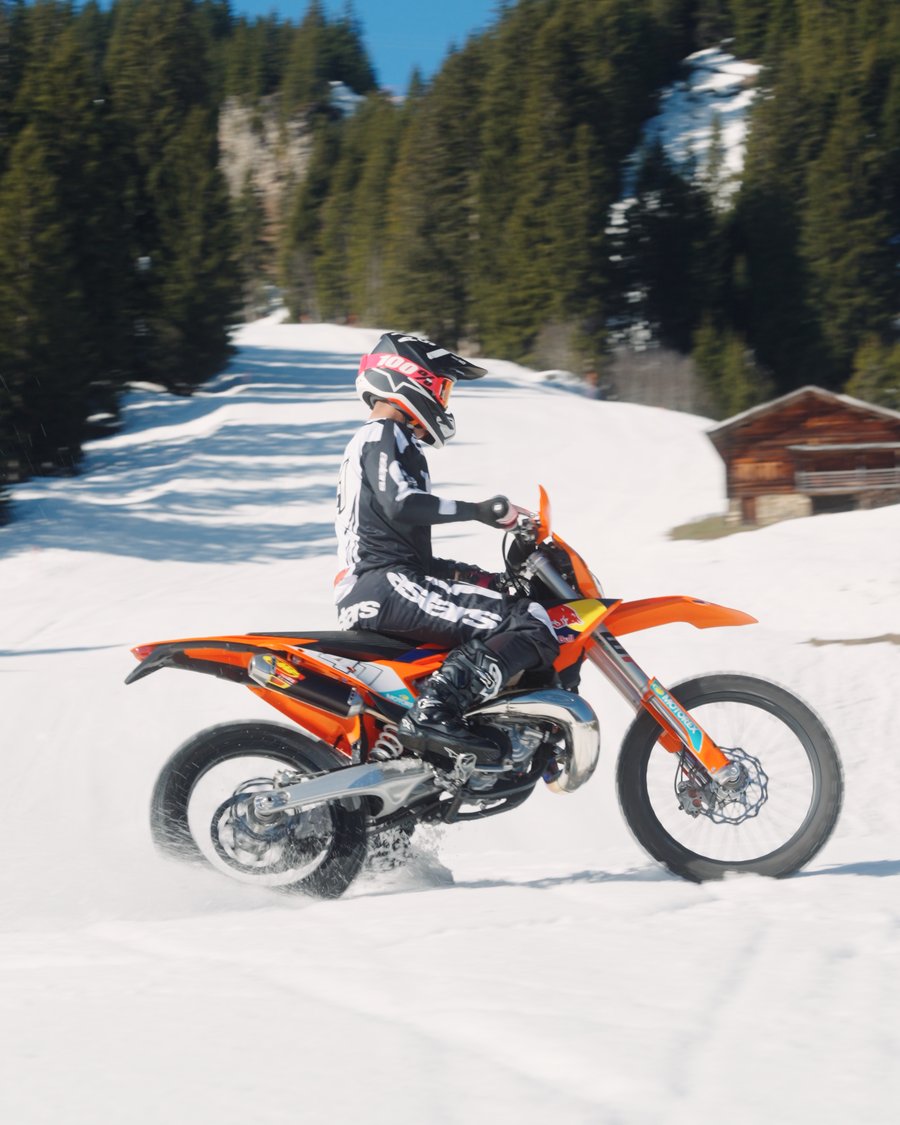 Fahrer auf einer KTM driftet durch den Schnee, Alphütte und Bergwald im Hintergrund