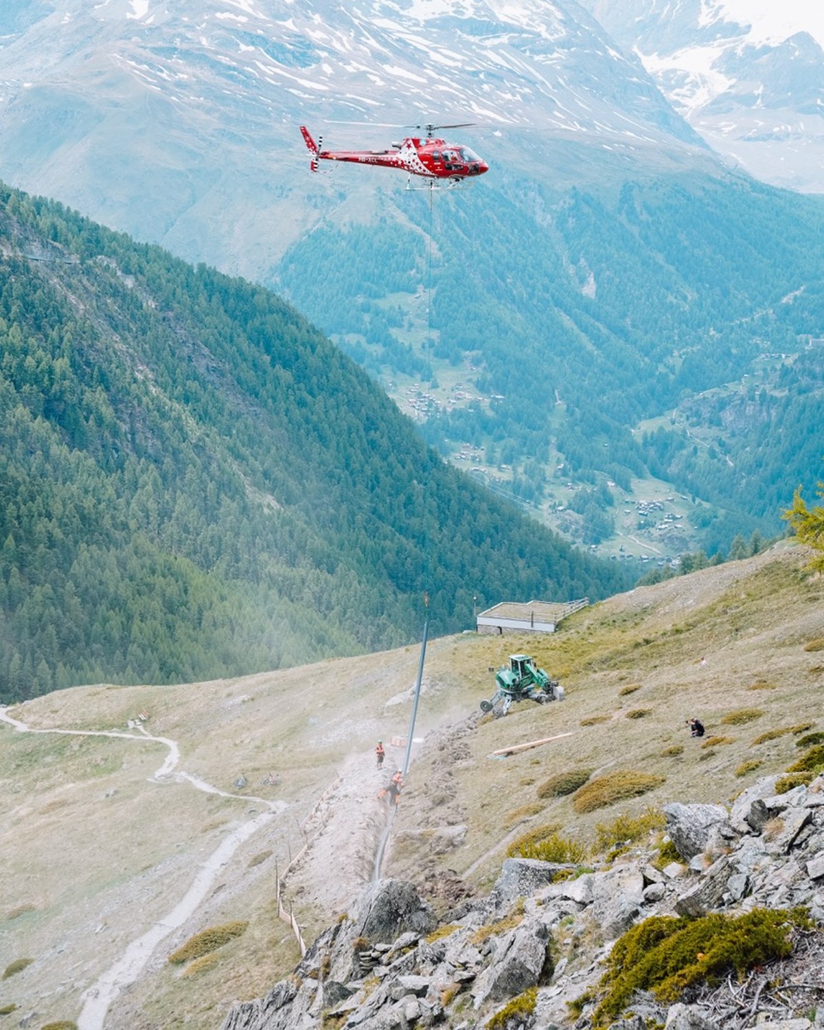 Helikopter transportiert eine Rohrleitung in einen Graben oberhalb von Zermatt, Bauteam und Bagger am Hang, Zermatt im Tal