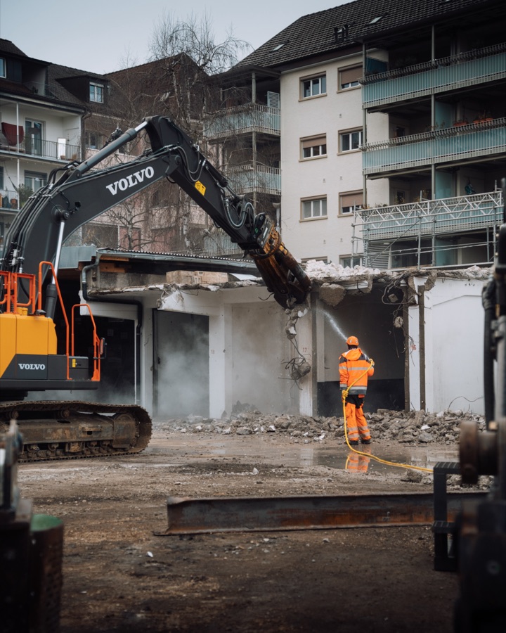 Abbruchbagger reisst ein Gebäude ein, Arbeiter kühlt mit Wasser