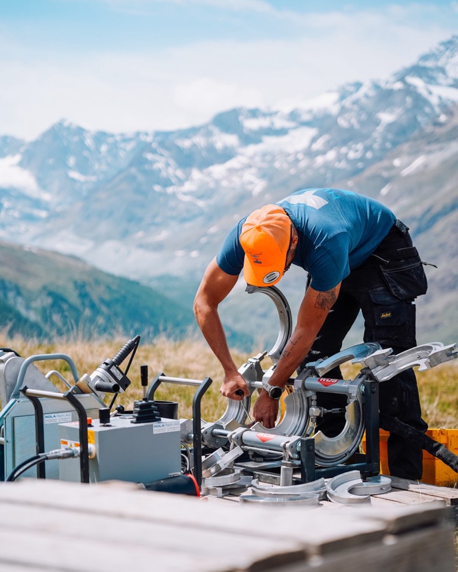 Mitarbeiter bedient die Rohrschweissmaschine, Alpenpanorama im Hintergrund